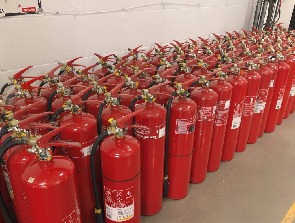 Multiple rows of red, decommissioned fire extinguishers grouped together on a facility floor, staged for collection and chemical neutralisation