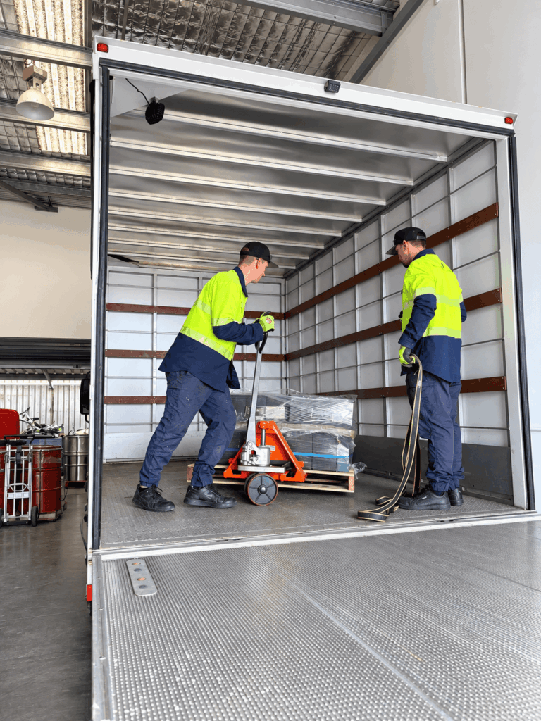 Two operators in high-visibility uniforms using a manual pallet jack to safely load a shrink-wrapped pallet into the back of a commercial transport truck.